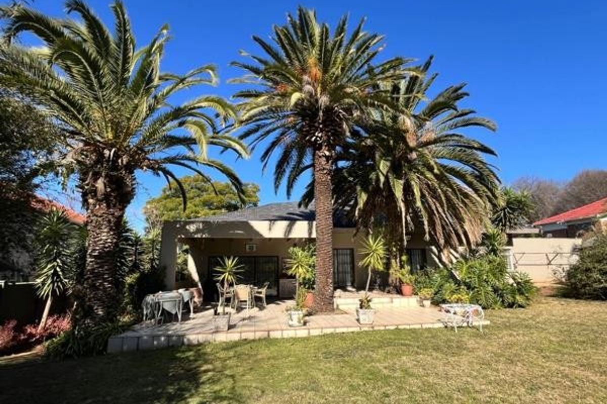 A rear view of the property featuring a covered tiled patio area overlooking a spacious garden with mature palm trees.