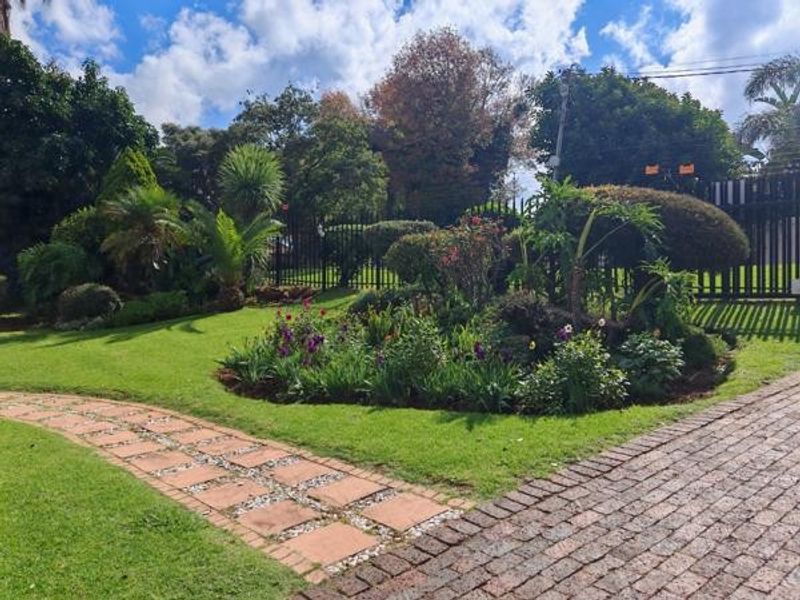 A well-maintained front garden featuring a paved walkway, brick driveway, and established landscaping enclosed by metal perimeter fencing.