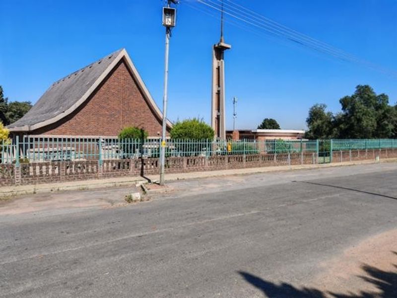Street view of a commercial property featuring a distinctive brick building with a steep pitched roof, concrete tower, and secure perimeter fencing.