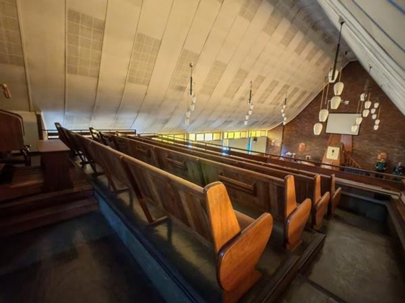 Interior view of a commercial assembly hall or chapel featuring tiered wooden pews, exposed brickwork, and a high sloped ceiling with pendant lighting.
