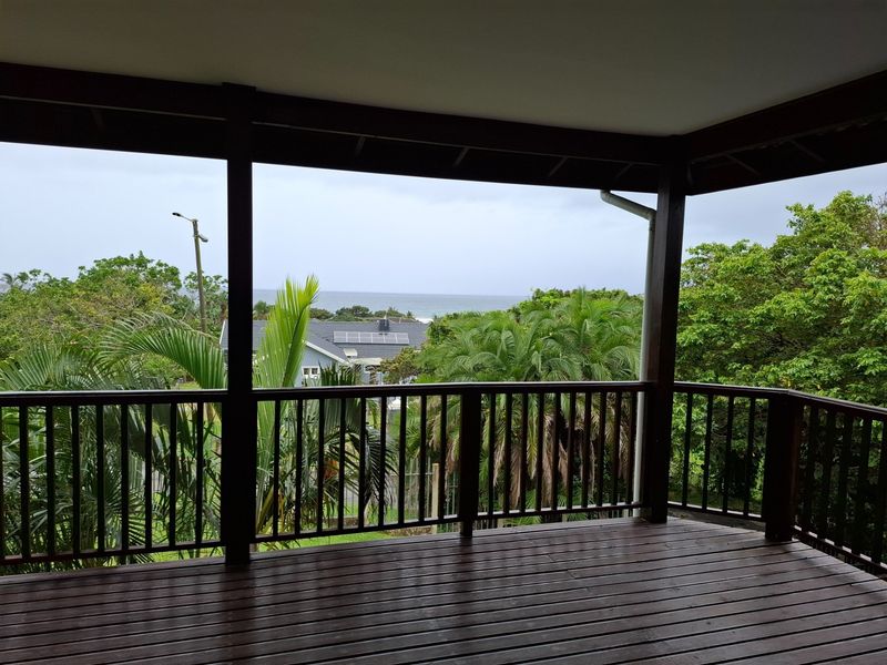 A spacious covered timber balcony featuring a wooden balustrade and offering scenic views of the surrounding greenery and distant ocean.