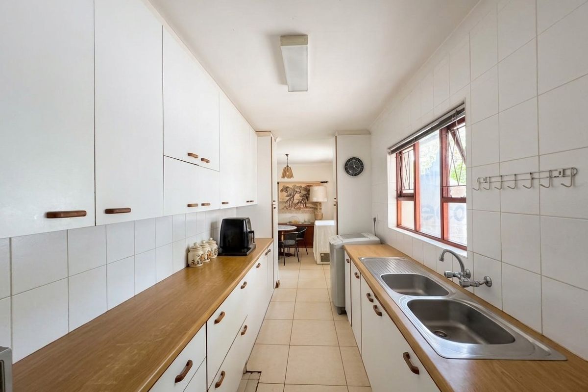A functional galley-style kitchen featuring white cabinetry, wooden countertops, a double stainless steel sink, and tiled flooring with a view into the dining area.