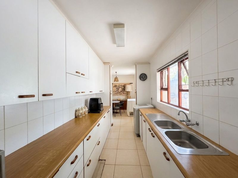 A functional galley-style kitchen featuring white cabinetry, wooden countertops, a double stainless steel sink, and tiled flooring with a view into the dining area.