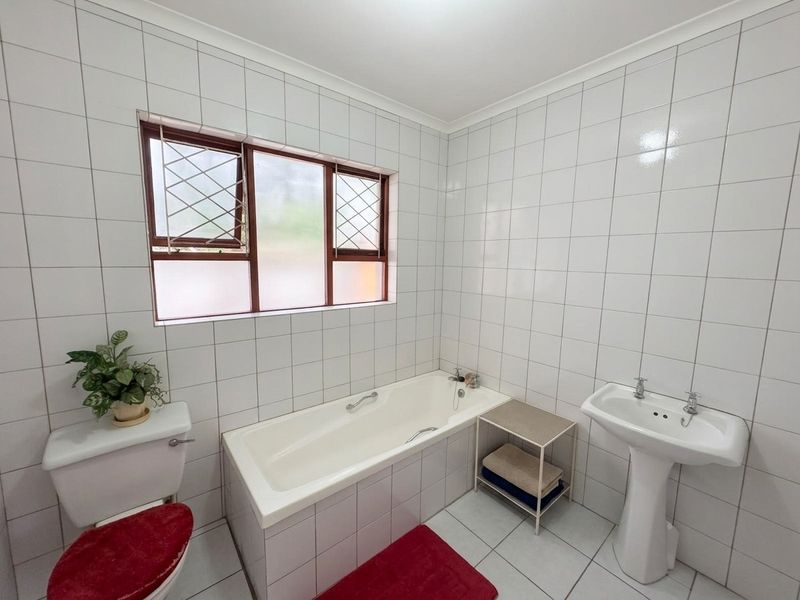 A functional bathroom featuring a pedestal basin, built-in bathtub, and full-height white ceramic wall tiling with a window for natural light.