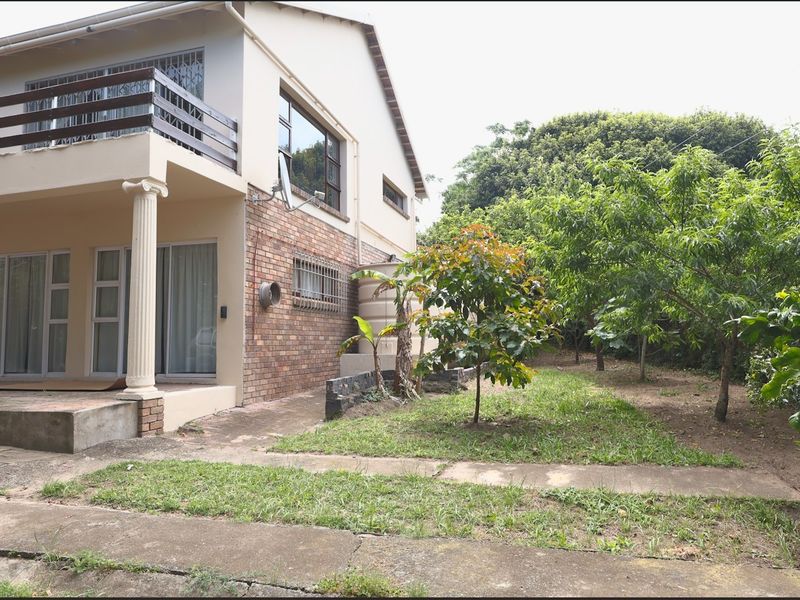 Rear view of a two-story house featuring a brick facade, a balcony, and a concrete patio area leading into a garden with mature trees.