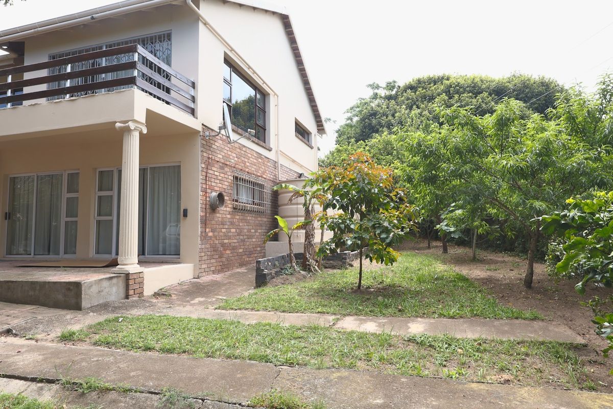 Rear view of a two-story house featuring a brick facade, a balcony, and a concrete patio area leading into a garden with mature trees.