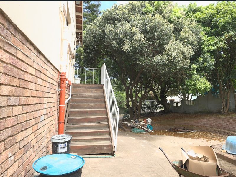 Rear exterior view featuring a brick facade, timber staircase with metal railing, and a paved patio area with mature trees.