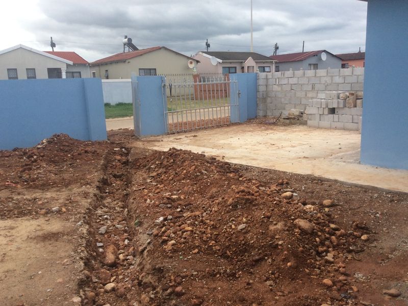 Front exterior view showing an unfinished driveway area with exposed soil, boundary walls, and a metal gate under construction.