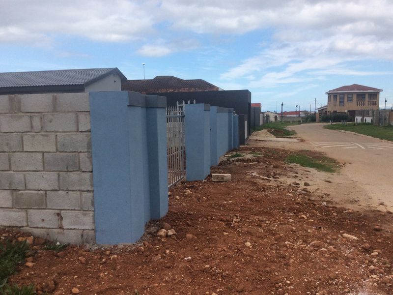 A street-level view showing a partially completed boundary wall with metal gate and unpaved ground along the roadside.