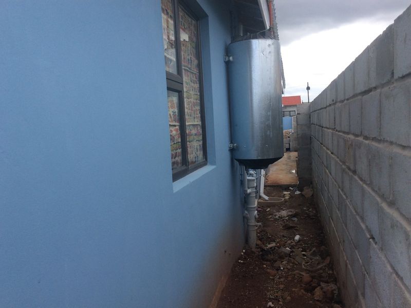 Side exterior view showing an external geyser installation, aluminium window frame, and concrete block boundary wall with unpaved ground.