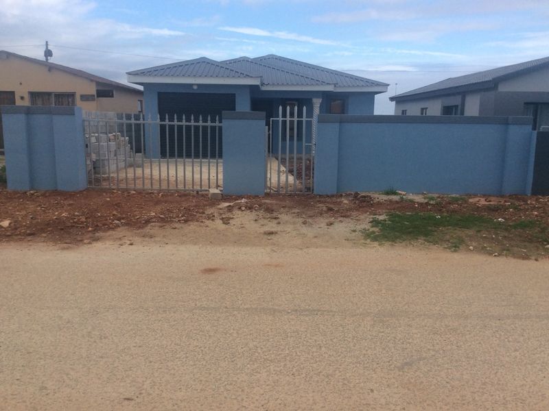Front view of a single-story house featuring a blue exterior, metal perimeter fencing, and a gated entrance with an unpaved driveway area.