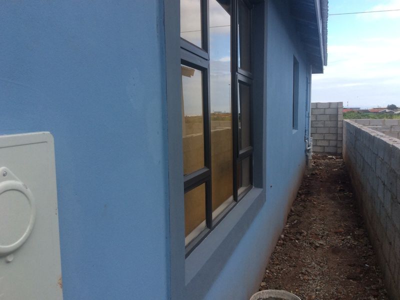 Side view of the property showing blue plastered exterior walls, dark aluminium window frames, and an unfinished ground area with concrete block boundary walls.