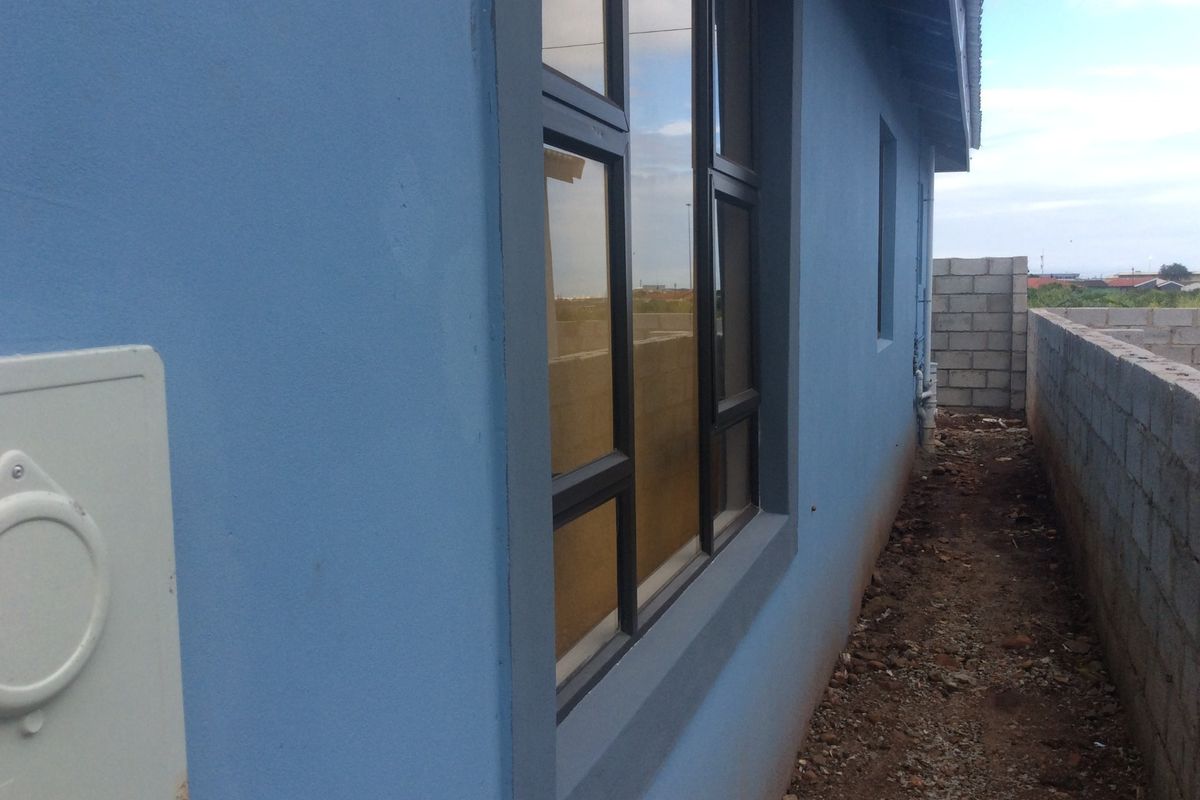 Side view of the property showing blue plastered exterior walls, dark aluminium window frames, and an unfinished ground area with concrete block boundary walls.