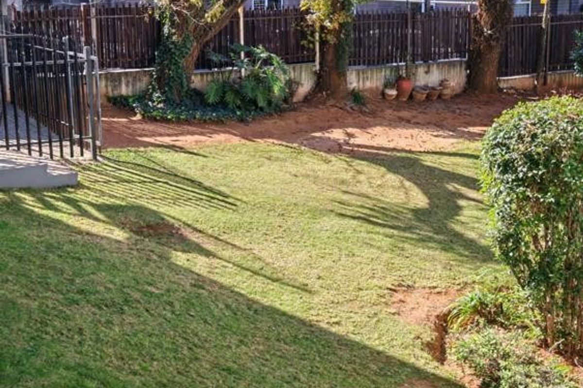A private garden area featuring a lawn, established trees, and metal perimeter fencing, providing a functional outdoor space for the apartment.
