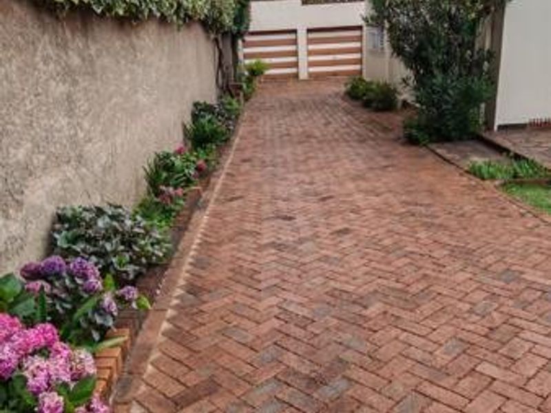 A well-maintained brick paved driveway leading to a double garage, flanked by a boundary wall and established garden beds.