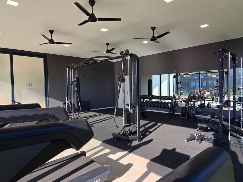 A modern, well-equipped gym featuring rubberized flooring, large wall mirrors, recessed lighting, ceiling fans, and expansive sliding glass doors.