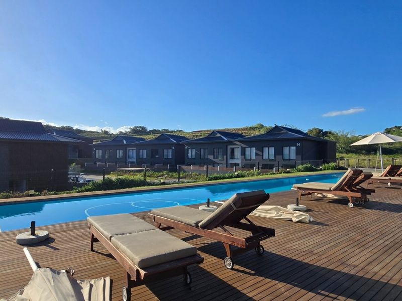 A spacious outdoor pool area featuring a long swimming pool set within a large timber deck, overlooking a landscaped estate with modern residential units.