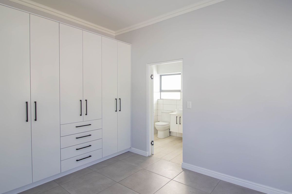 A clean, modern bedroom featuring extensive built-in white cabinetry, neutral tiled flooring, and direct access to an en-suite bathroom.
