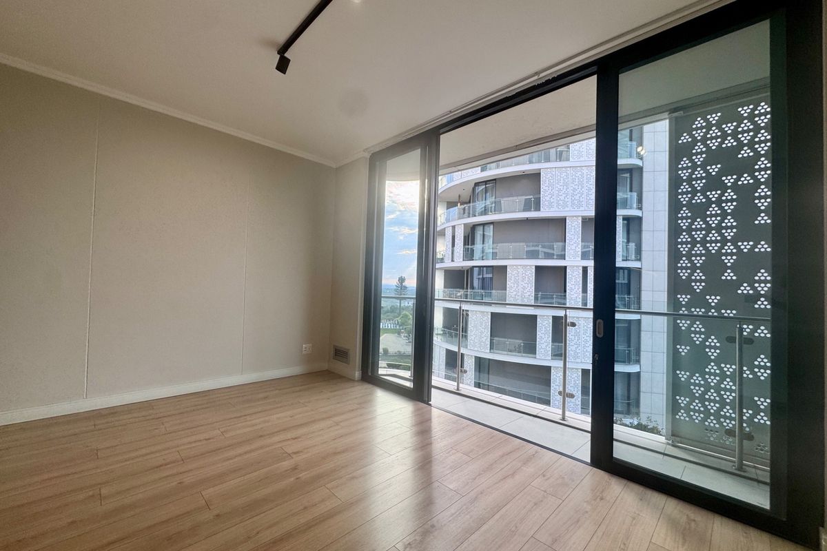 A bright, empty bedroom featuring modern laminate flooring, track lighting, and large sliding glass doors that open onto a private balcony.