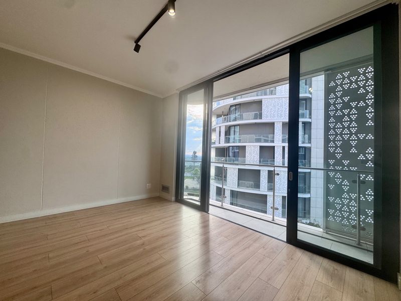 A bright, empty bedroom featuring modern laminate flooring, track lighting, and large sliding glass doors that open onto a private balcony.