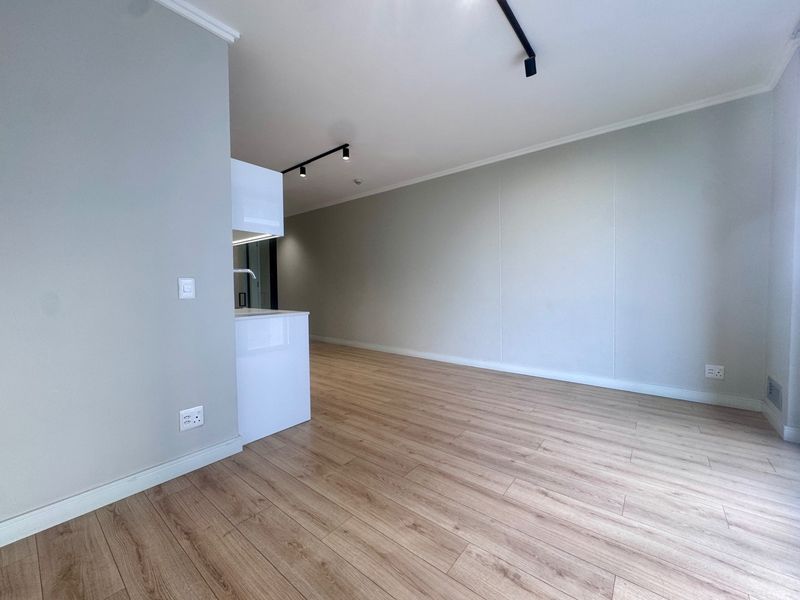 A bright, modern open-plan living area featuring light wood-look laminate flooring, neutral walls, and contemporary track lighting with a compact kitchen section.
