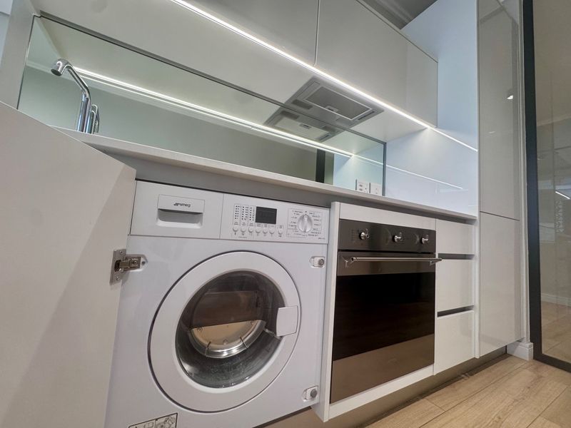 Modern kitchen featuring handleless white cabinetry, integrated washing machine, built-in oven, and a sleek mirror splashback with integrated LED lighting.