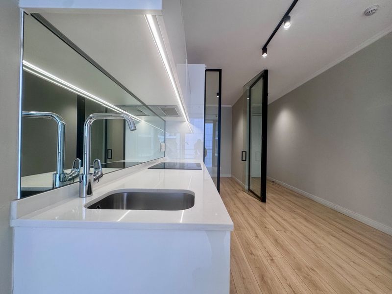Modern kitchen featuring sleek white cabinetry, quartz countertops, integrated hob, and mirrored splashback, leading into an open-plan living area with contemporary glass doors.