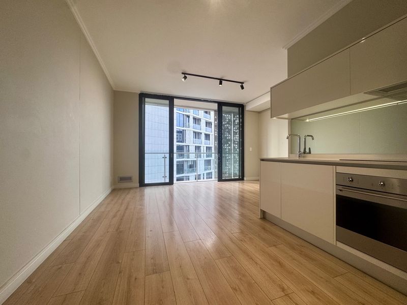 Modern open-plan living area featuring light laminate flooring, sleek built-in kitchen cabinetry with integrated oven, and large sliding doors leading to a private balcony.