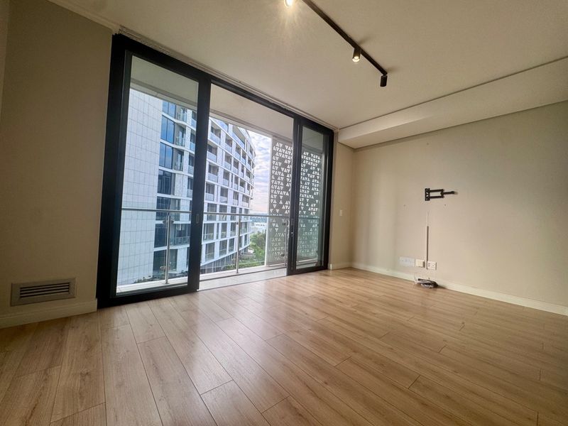 A bright, empty living room featuring modern laminate flooring, track lighting, and large sliding glass doors that open onto a balcony with urban views.
