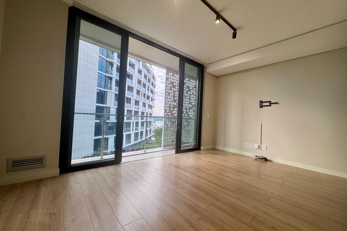 A bright, empty living room featuring modern laminate flooring, track lighting, and large sliding glass doors that open onto a balcony with urban views.