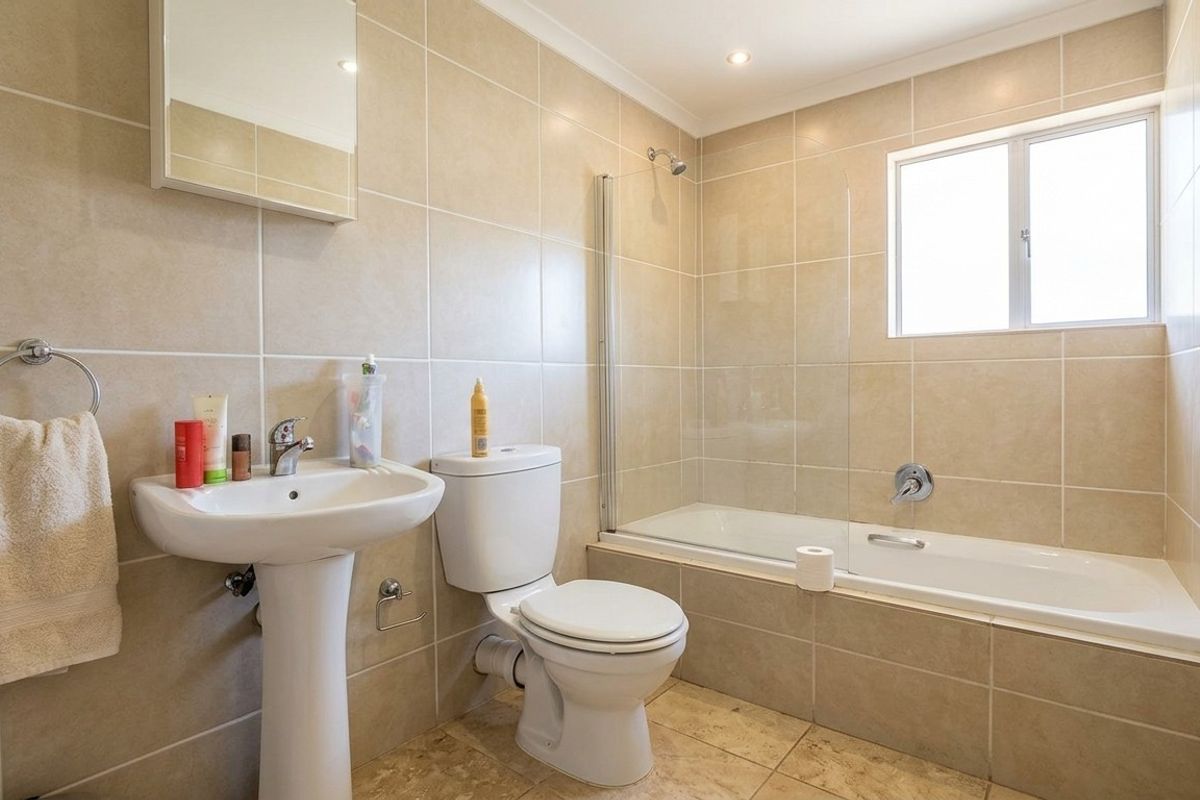 A well-maintained bathroom featuring a pedestal basin, toilet, and a bathtub with a glass shower screen, finished with neutral wall and floor tiling.