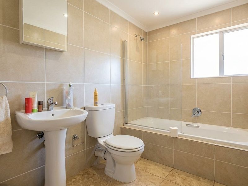 A well-maintained bathroom featuring a pedestal basin, toilet, and a bathtub with a glass shower screen, finished with neutral wall and floor tiling.