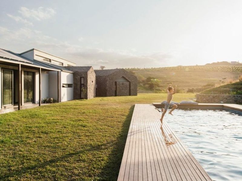 A modern outdoor pool area featuring extensive timber decking, stone-clad building structures, and a well-maintained lawn with scenic landscape views.