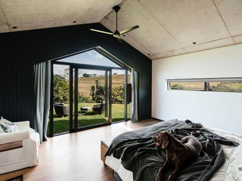 Modern bedroom featuring an exposed concrete ceiling, timber flooring, and large glass doors opening to a scenic rural view.