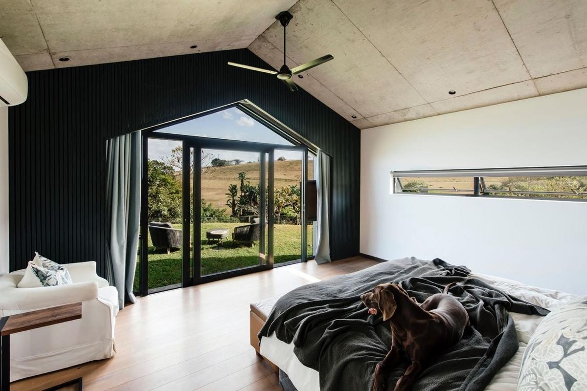 Modern bedroom featuring an exposed concrete ceiling, timber flooring, and large glass doors opening to a scenic rural view.