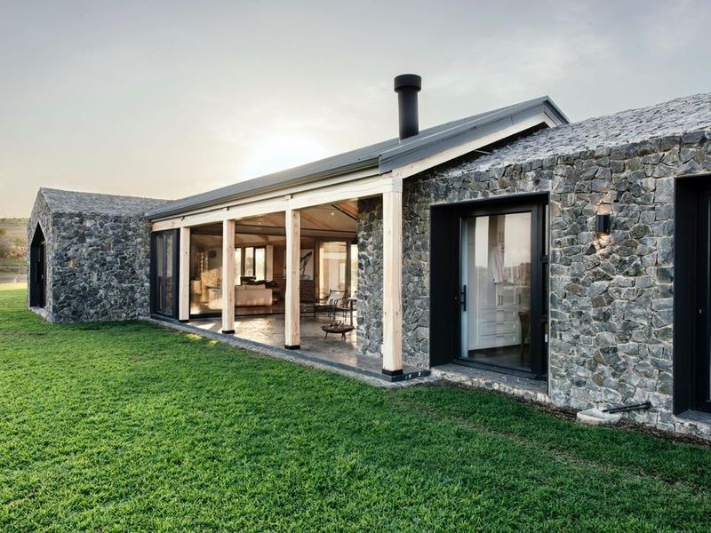 A modern architectural home featuring natural stone cladding, a covered patio with timber posts, and expansive sliding glass doors overlooking a manicured lawn.