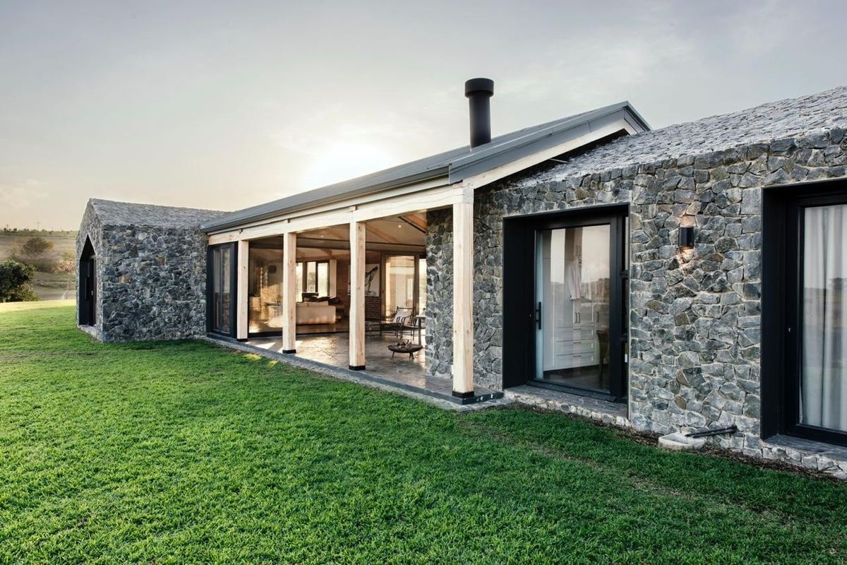 A modern architectural home featuring natural stone cladding, a covered patio with timber posts, and expansive sliding glass doors overlooking a manicured lawn.