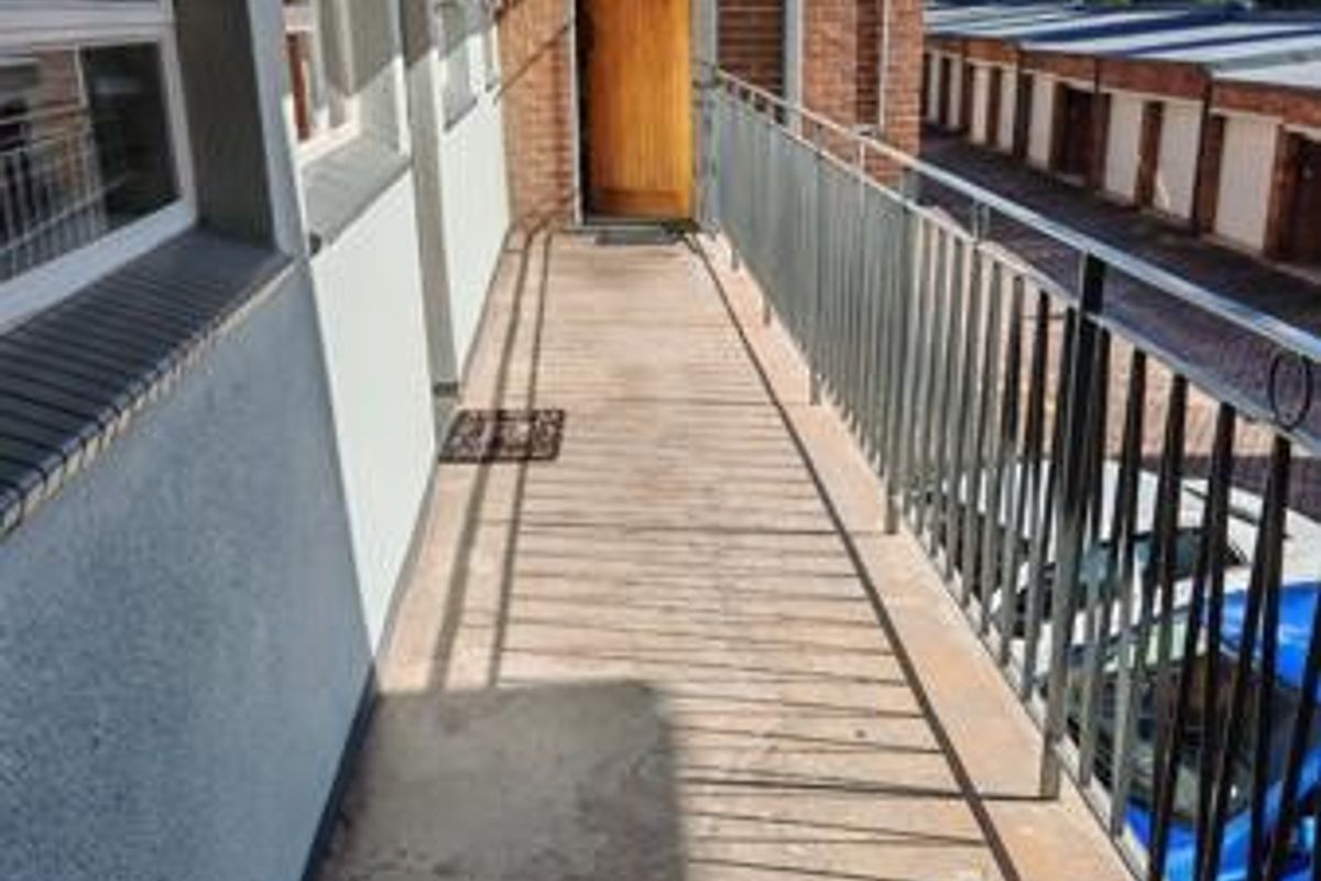 An open-air apartment balcony featuring a metal balustrade, brick exterior wall, and concrete flooring, providing access to the main entrance door.