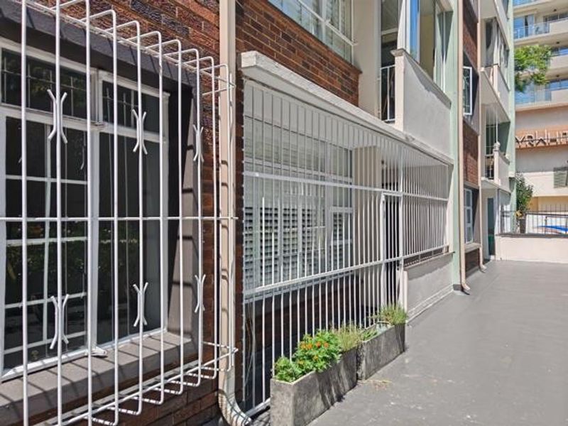 Exterior view of an apartment building facade featuring brickwork, protective security bars on windows, and integrated concrete planters along the walkway.