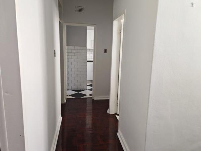 A clean, empty hallway featuring dark parquet flooring and white walls, providing access to adjacent rooms including a kitchen with tiled splashbacks.