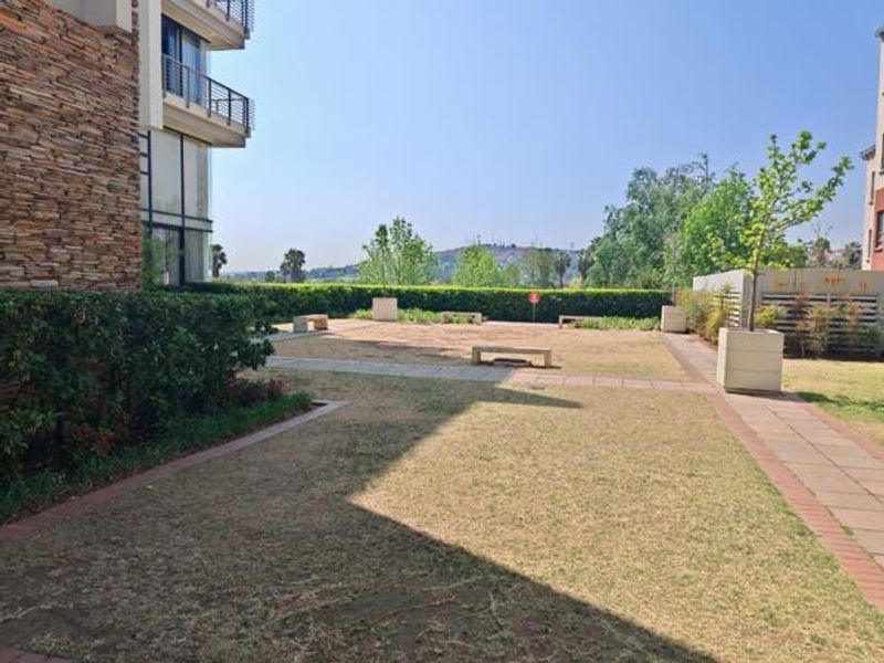 A communal garden area featuring paved walkways, stone-clad building exterior, and established hedging with views of the surrounding landscape.