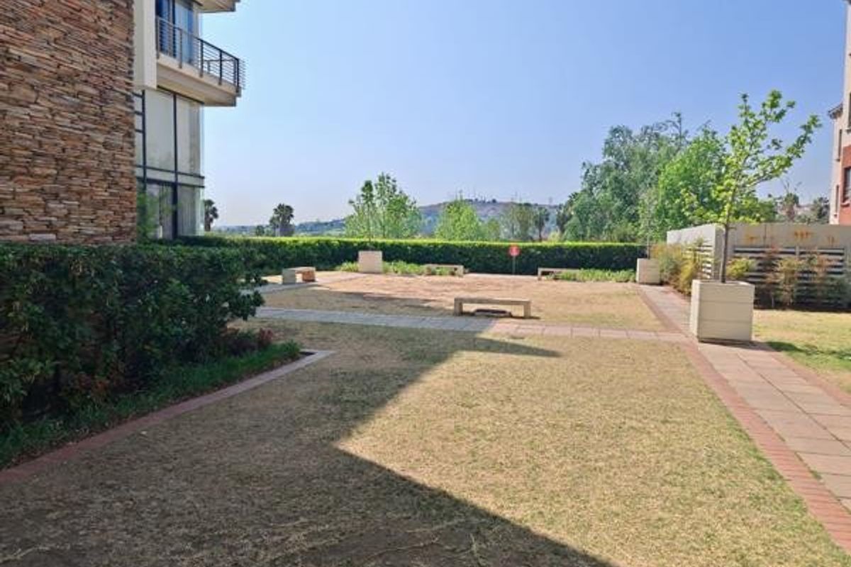 A communal garden area featuring paved walkways, stone-clad building exterior, and established hedging with views of the surrounding landscape.