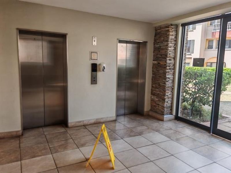 A modern building entrance lobby featuring dual stainless steel elevators, decorative stone cladding, and large glass doors providing natural light and garden views.