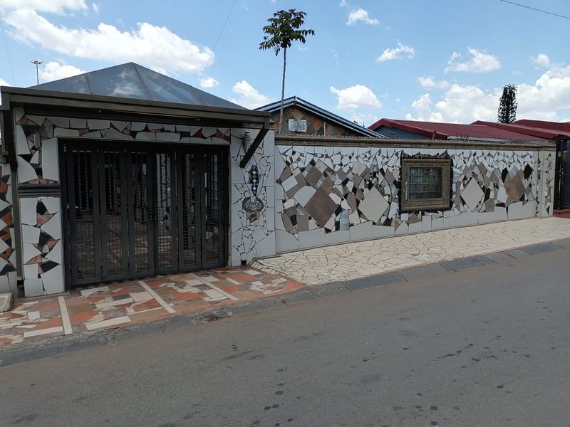 Front exterior view of the property featuring a distinctive mosaic-tiled boundary wall, metal security gate, and paved driveway area.
