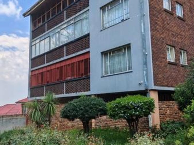 A multi-story commercial building featuring a brick facade, enclosed balconies with security bars, and established garden landscaping at the front.