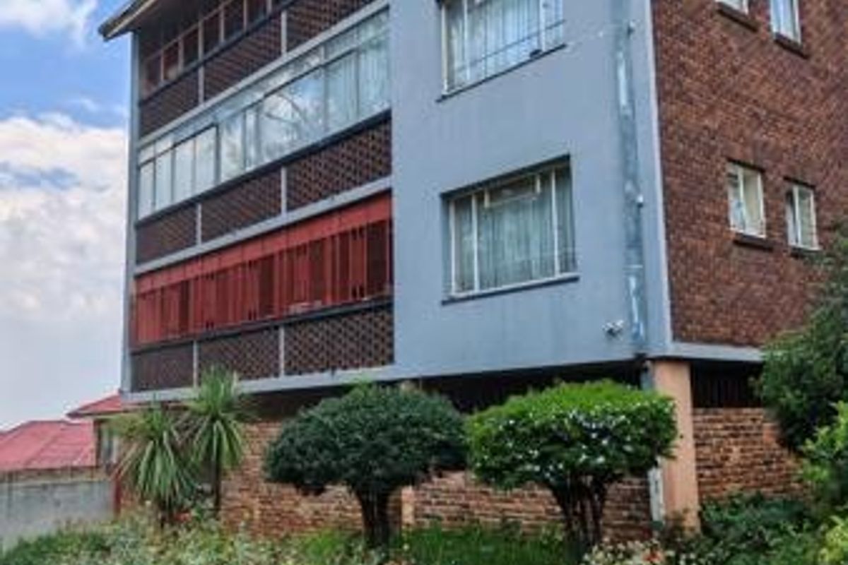 A multi-story commercial building featuring a brick facade, enclosed balconies with security bars, and established garden landscaping at the front.