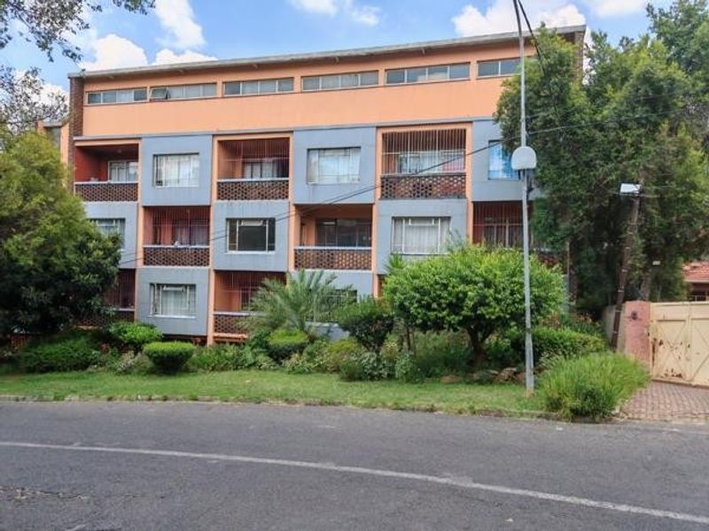 A multi-story commercial or residential building facade featuring balconies with security grilles, set behind a landscaped garden and street-level access.
