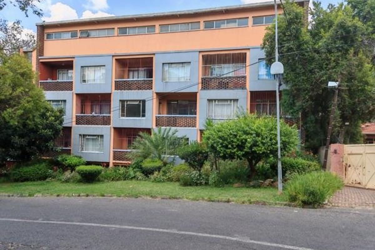 A multi-story commercial or residential building facade featuring balconies with security grilles, set behind a landscaped garden and street-level access.