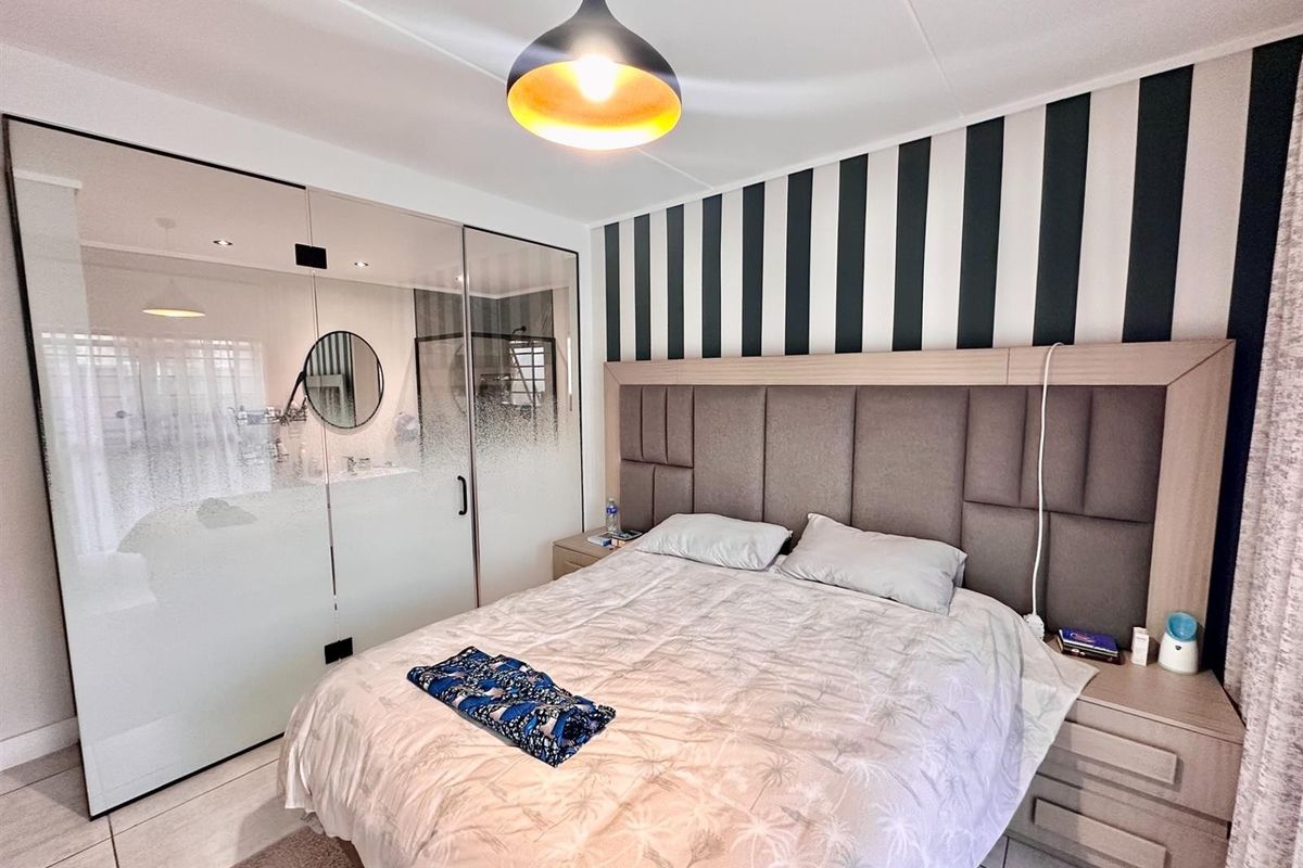 A modern bedroom featuring a built-in upholstered headboard, sleek glass partition wall, and contemporary pendant lighting with neutral tiled flooring.