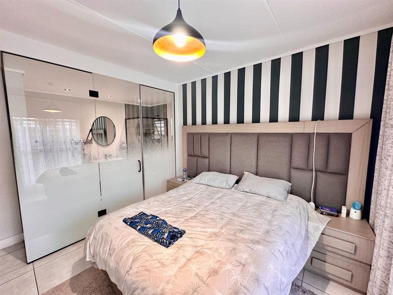 A modern bedroom featuring a built-in upholstered headboard, sleek glass partition wall, and contemporary pendant lighting with neutral tiled flooring.